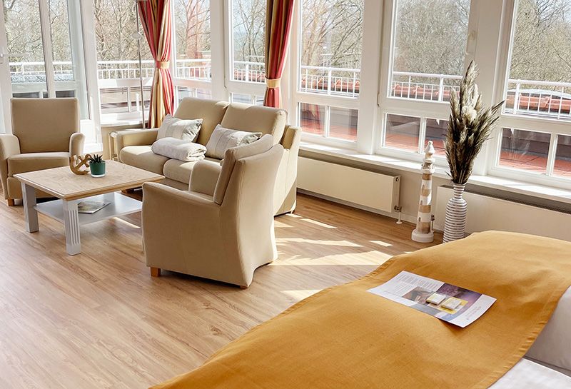 Suite mit Wohn- und Schlafbereich, Sitzecke mit Sofa, Sessel und Tisch, Doppelbett mit Tagesdecke und Prospekt, helle Fensterfront, umliegender Balkon mit Blick auf die Ostsee