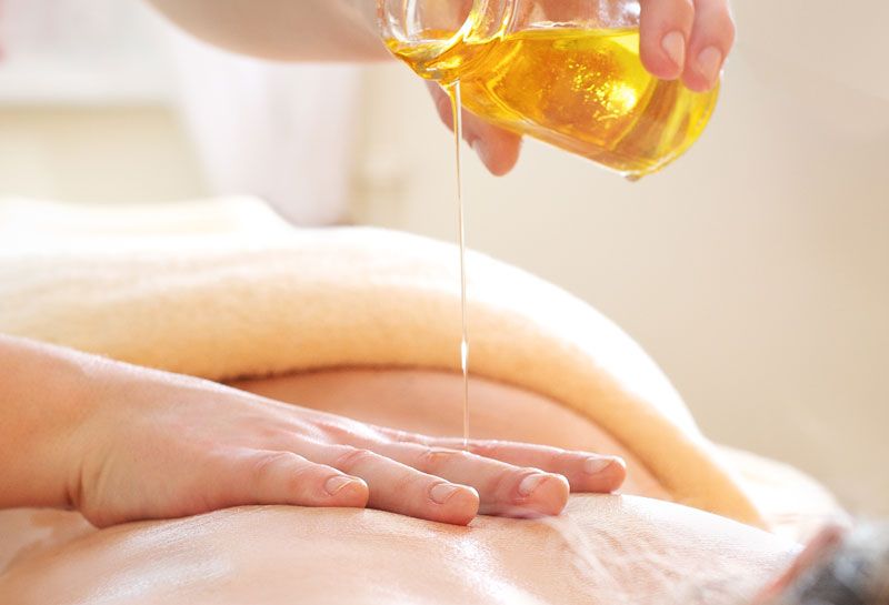 Körper-Rituale Hohe Wacht, Anschnitt vom Rücken, eine Hand hält ein Glasgefäß, in dem Galsgefäß ist Öl, massierende Frau gießt Öl auf Rücken, mit anderer Hand wird das Öl auf dem Rücken verteilt