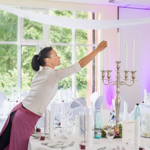 eingedeckter Banketttisch mit Geschirr, Besteck, Gläsern, Servietten, Buffet- und Getränkekarte, Mitte des Tisches silberner Kronleuchter Blumendeko, Frau mit weißer Bluse und weinroter Schürze zündet die Kerzen an, im Hintergrund Fensterfront