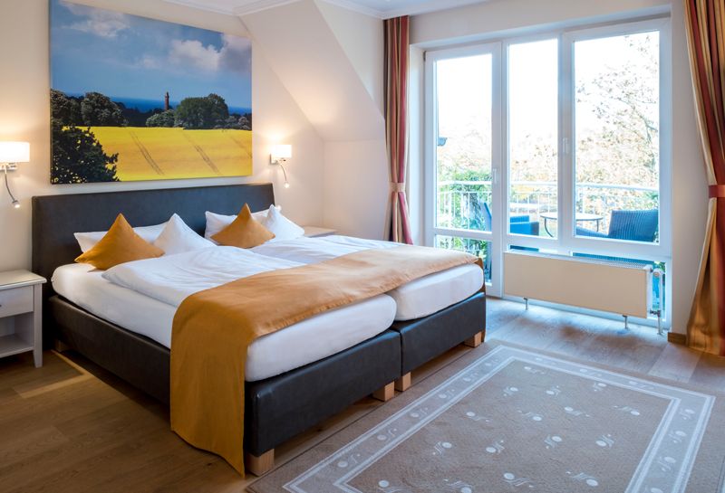 Schlafbereich im 1-Zimmer-Komfortappartement mit Schlafzimmer, an der Wand Leinwand mit Rapsmotiv, links vom Bett Fenster mit Terrassentür zum Balkon mit Sitzmöglichkeiten und kleinem Tisch, vor dem Bett liegt ein Teppich