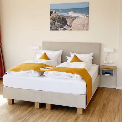Schlafbereich Ostsee-Suite mit Boxspringbett, über dem Kopfende Leinwand mit Strandmotiv, links und rechts neben dem Bett Lampen und Ablageflächen