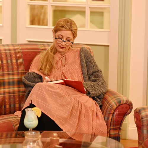 Bibliothek Hotel Hohe Wacht, Frau mit Brille sitzend auf einem karierten Sofa, Beine überschlagen, auf dem Schoß ein Buch, in der Hand ein Stift, im Vordergrund ein Glastisch, auf dem Tisch steht ein Cocktail