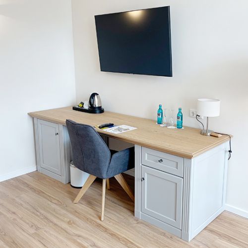Doppelzimmer Schreibtisch mit Stuhl an der Wand, über dem Schreibtisch ein TV-Gerät, auf dem Schreibtisch Lampe, Welcome Tray Kaffee/Tee, Wasserkocher, Minibar, Fernbedienung TV, Prospekt